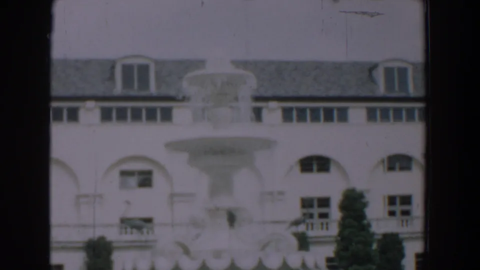 1957 Florida home-movie frame showing a large white building with arched windows, roof dormers, a fountain, and shrubs.