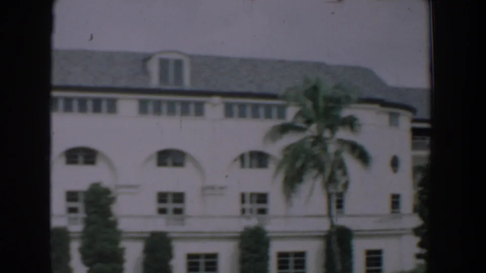 1957 Florida exterior view of a long white building with arched windows, dormer roof details, palm trees, and manicured greenery.