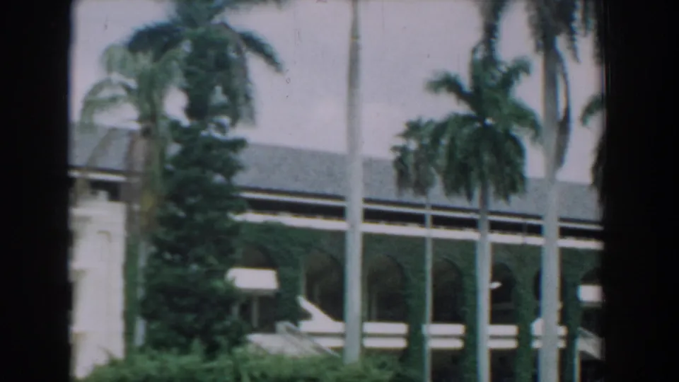 1957 Florida home-movie frame showing palms, hedges, and a white building section with arched lower openings and shaded facade details.