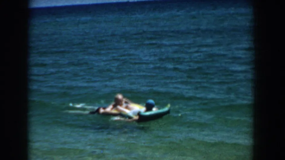 Two people continue floating on an inflatable raft in Florida ocean water, seen through soft 1957 8mm color footage.