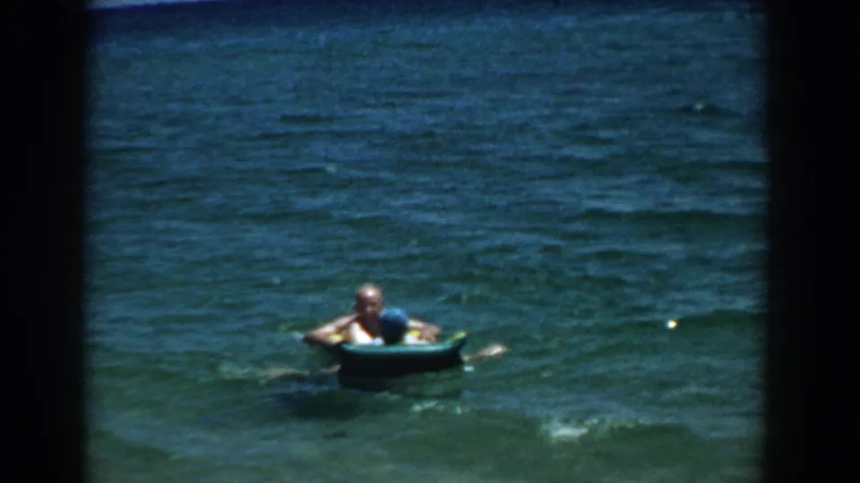 Home-movie frame of two people beside a small blue raft, with textured lake water filling most of the view.