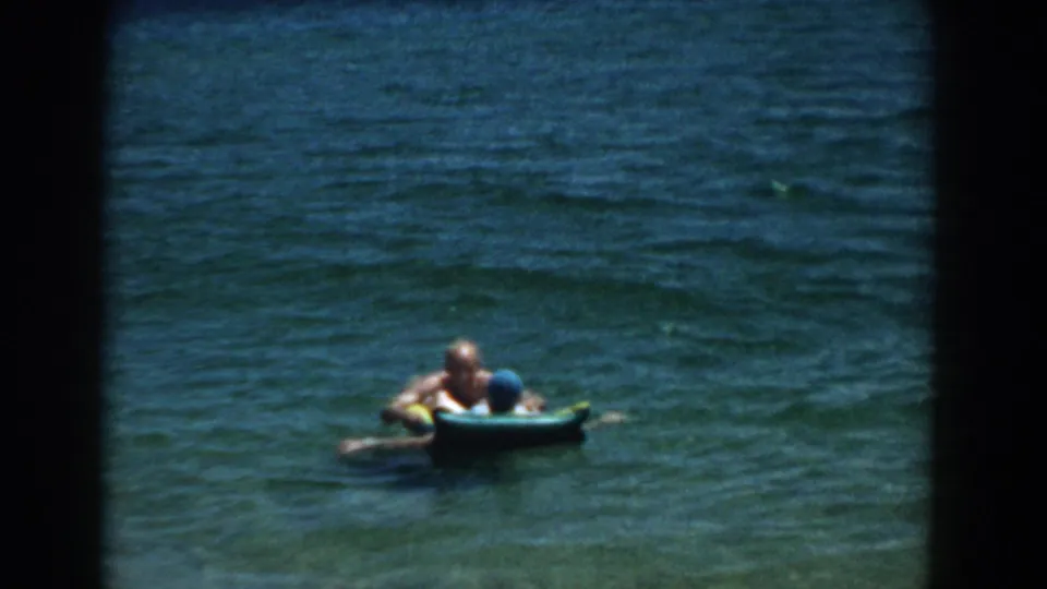 A 1957 Florida lake frame shows two people partly behind a blue inflatable raft, surrounded by blue-green rippling water.