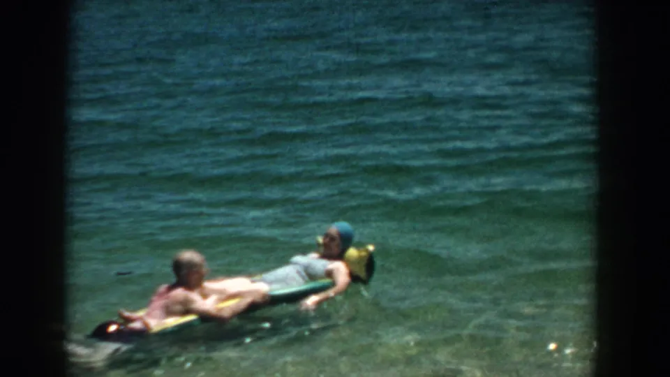 1957 Florida home-movie frame of an adult couple reclining on a green float, with the woman wearing a blue swim cap.