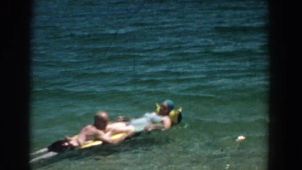 1957 Florida 8mm frame showing the couple on an inflatable float, with the woman in a blue swim cap and water rippling around them.