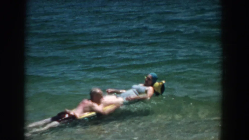 1957 Florida home-movie frame of an adult couple drifting on a float in clear blue-green water, with soft 8mm color and grain.