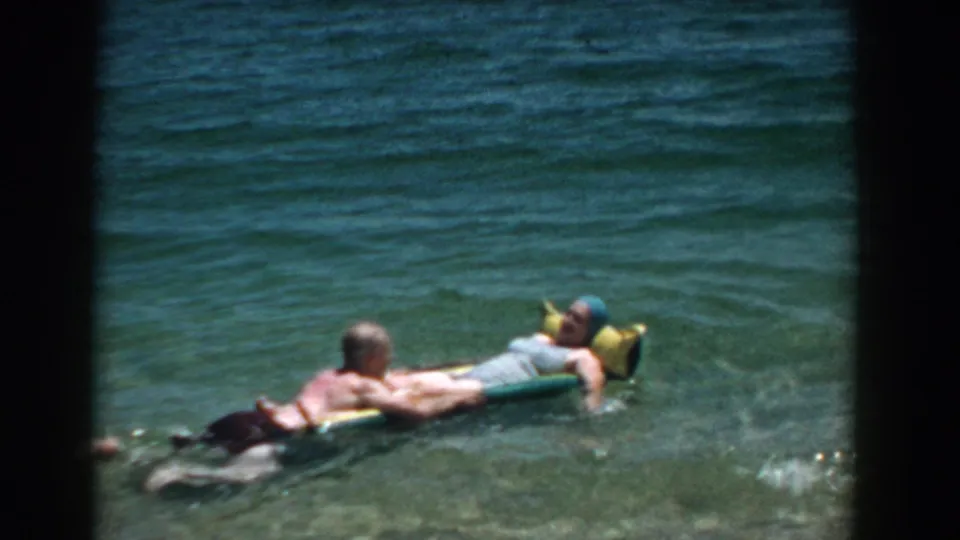1957 Florida 8mm frame showing two adults relaxing on a yellow and green inflatable raft in clear beach water.