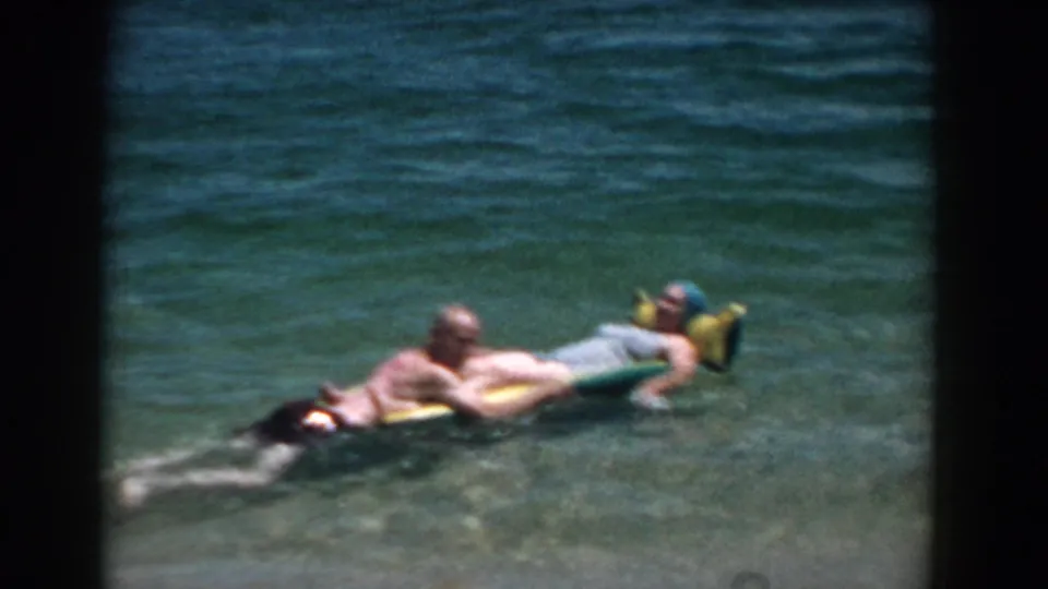 1957 Florida beach frame with two adults stretched across a yellow and green raft in shallow turquoise water.