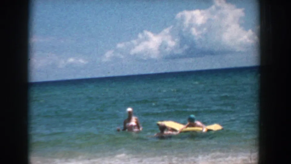1957 Florida 8mm frame showing adults in shallow surf near an inflatable raft, with open ocean and clouds in the background.