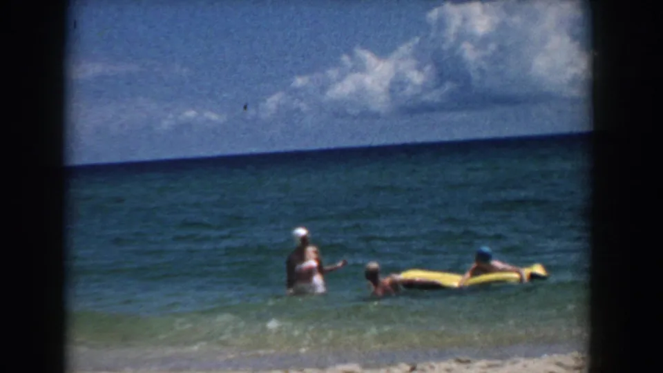1957 Florida beach frame with adults in the surf next to a yellow raft, ocean horizon, and bright clouds.