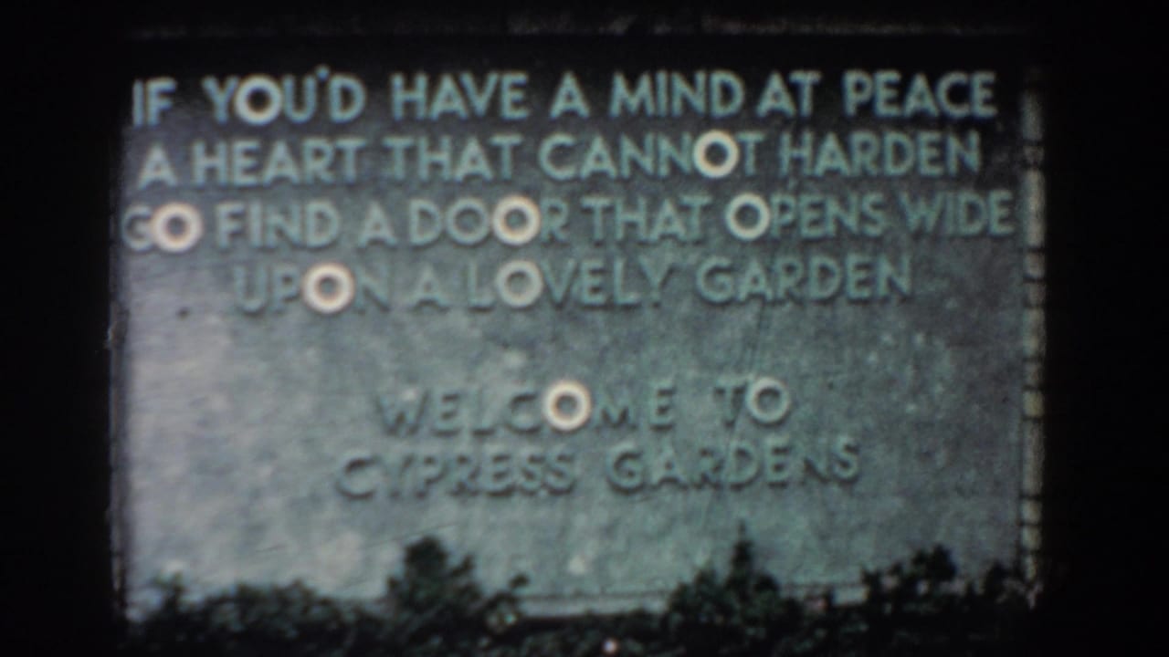 1957 Florida 8mm frame showing a weathered stone Cypress Gardens welcome sign with a carved garden poem and low foliage.