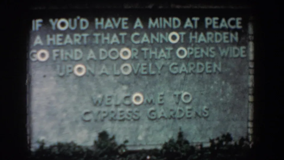 1957 Florida 8mm frame showing a weathered stone Cypress Gardens welcome sign with a carved garden poem and low foliage.