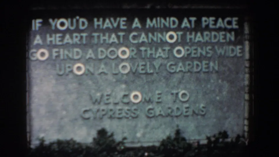1957 Florida frame of the Cypress Gardens welcome sign, with the engraved poem visible above low plants and dark film edges.