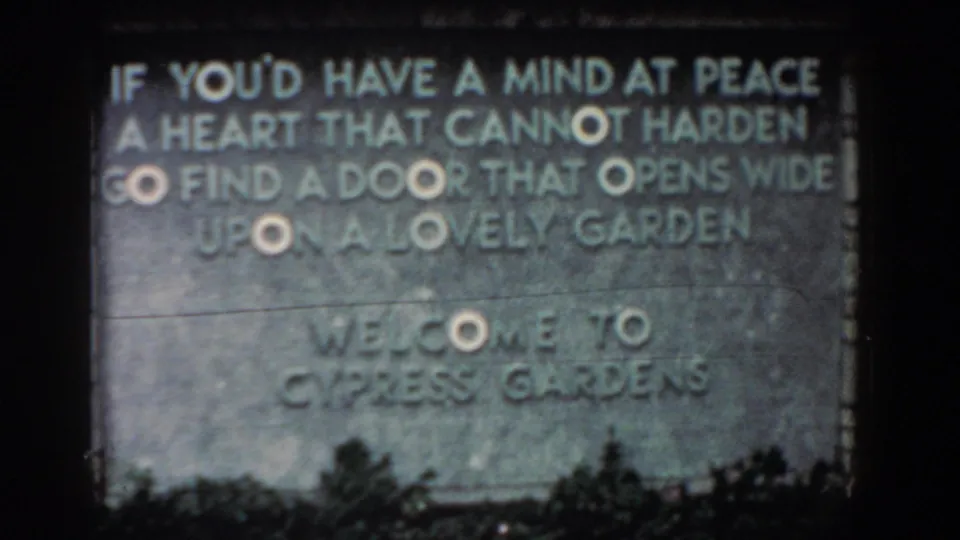 1957 Florida 8mm frame of the Cypress Gardens welcome sign, with carved text, low foliage, and a visible horizontal film artifact.