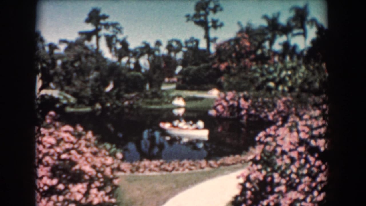 1957 Florida 8mm frame showing a white canoe on a small pond amid palms, pink shrubs, lawn, and a curving path.