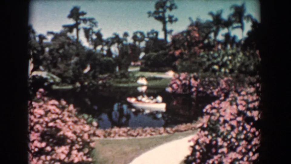 1957 Florida 8mm frame showing a white canoe on a small pond amid palms, pink shrubs, lawn, and a curving path.
