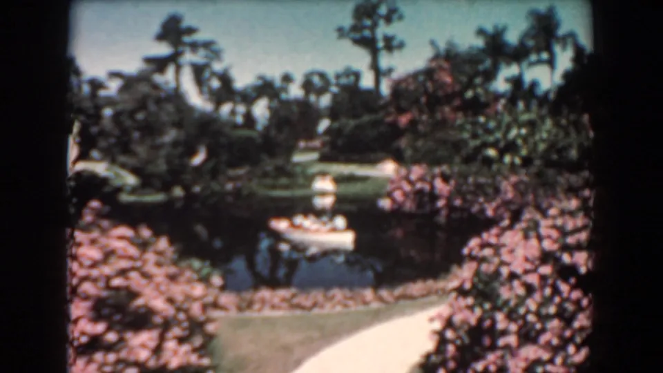 1957 Florida home-movie frame with a small white canoe crossing a pond below tall palms and dense flowering plantings.