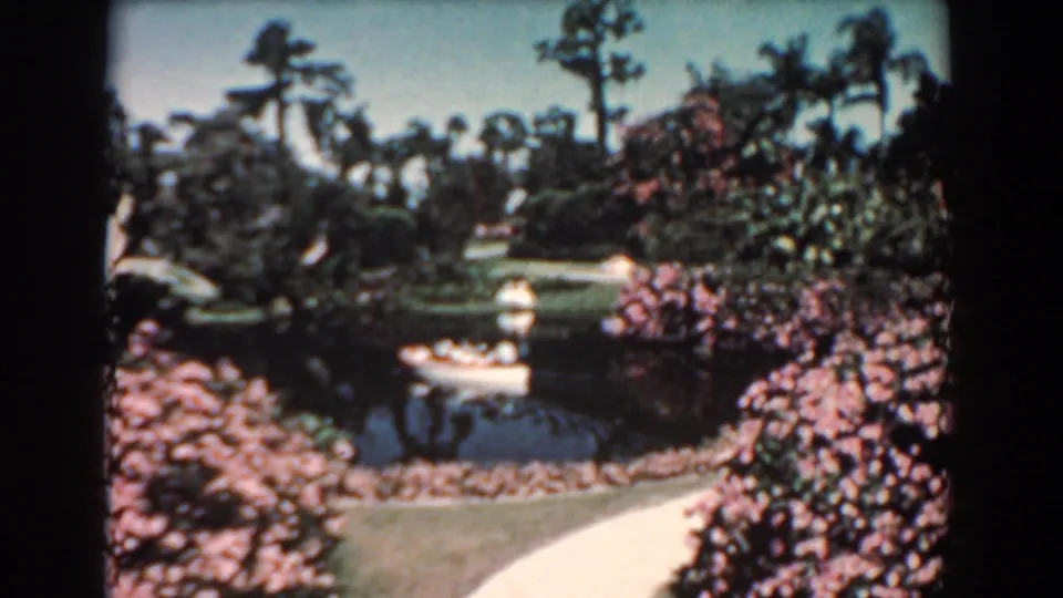 1957 Florida 8mm still showing canoe passengers on a quiet pond, with palm trees, shrubs, lawn, and a path in view.