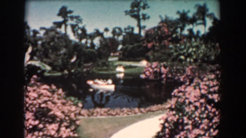1957 Florida home-movie frame of a white canoe on a landscaped park pond, bordered by palms, shrubs, grass, and a walkway.