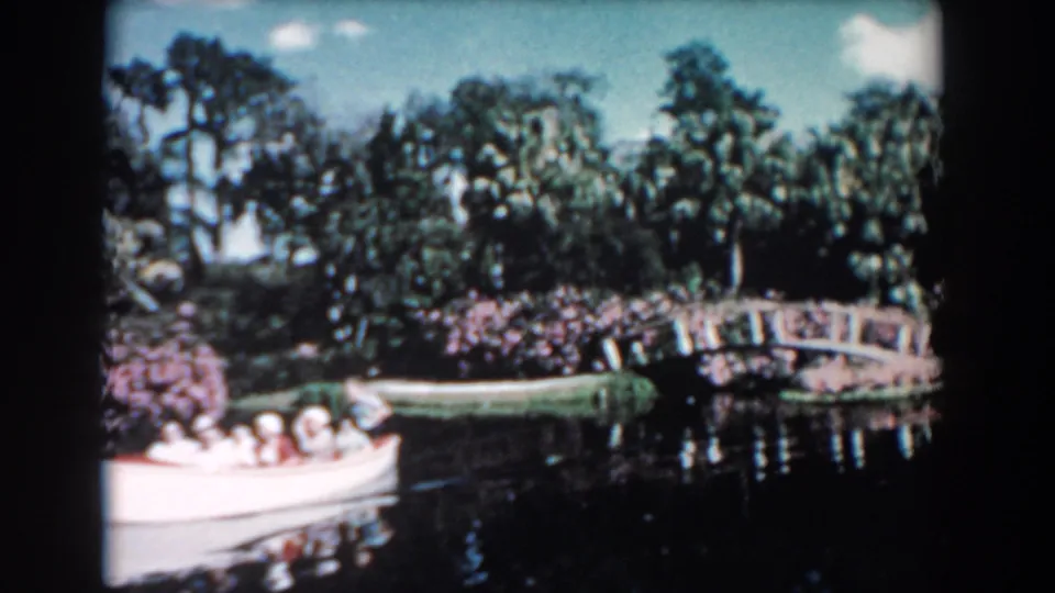 1957 Florida 8mm frame showing a white tour boat with passengers near an arched garden bridge, trees, shrubs, and calm water.