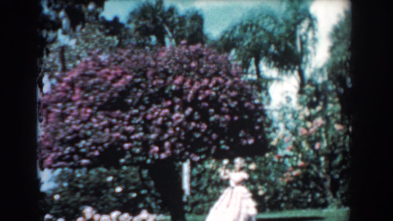 Woman in a pale long dress standing near a large purple flowering bush in a lush garden.