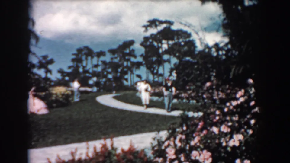 Visitors in 1957 Florida appear on a winding garden walk bordered by blooming shrubs, lawn, and tall tropical-looking trees.