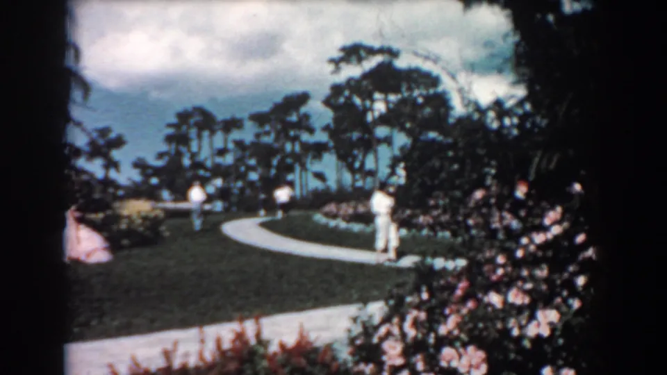 1957 Florida garden view with several adults near a curving walkway, landscaped lawn, shrubs, and tall trees under clouds.