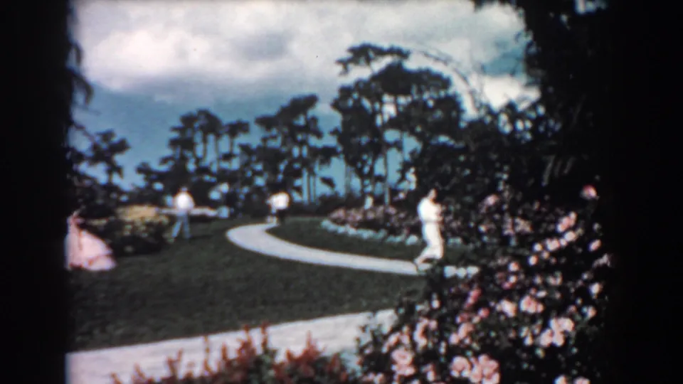Soft-focus 1957 Florida 8mm frame of adults on and near a garden walkway, with flowers, lawn, trees, and bright sky.