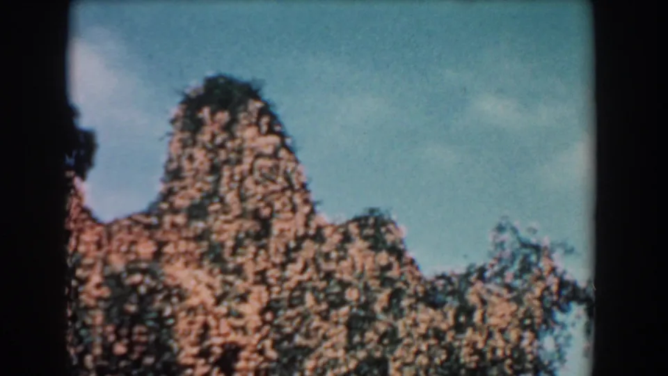 1957 Florida garden frame showing a rocky mound or planted rock feature with trees and blue sky in vintage 8mm color.