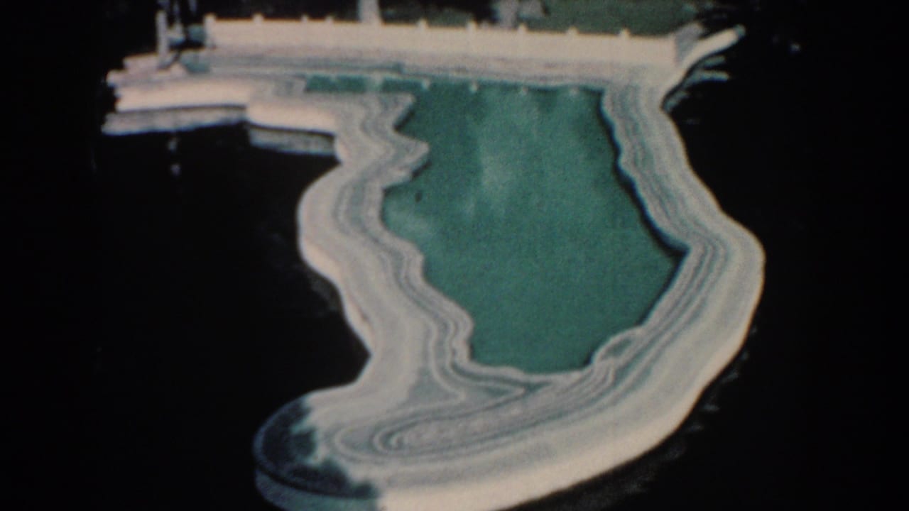 1957 Florida home-movie frame showing a shallow turquoise pool shaped like the state, with white concrete edges near a dock over dark water.