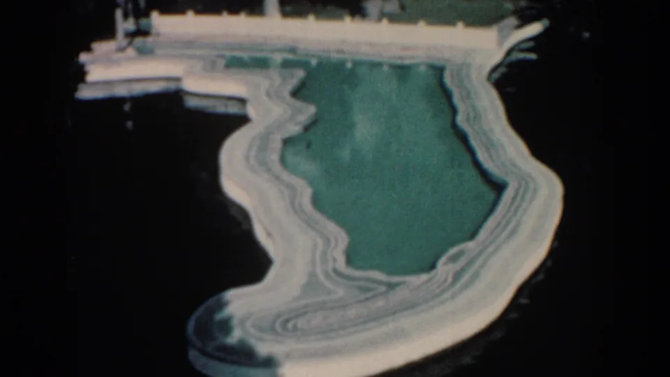 1957 Florida home-movie frame showing a shallow turquoise pool shaped like the state, with white concrete edges near a dock over dark water.