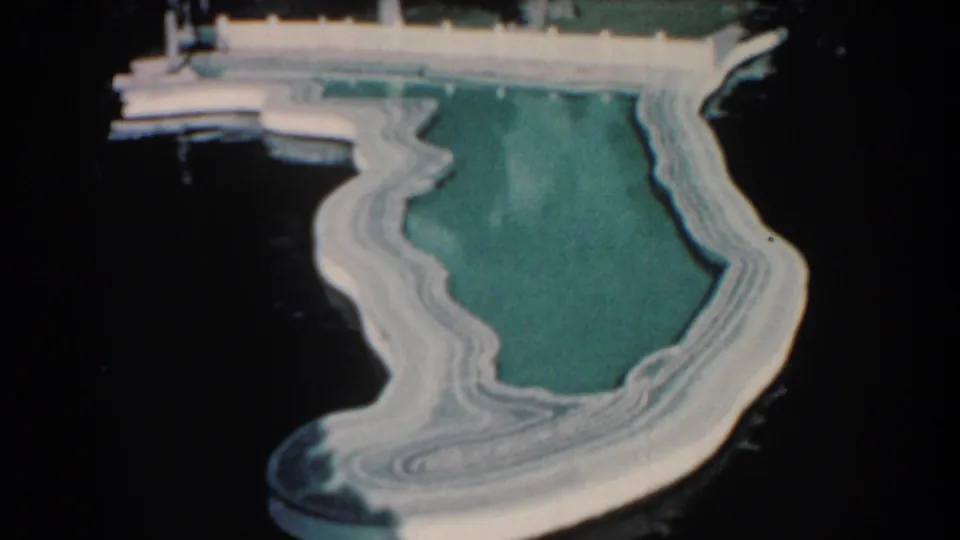 The Florida-shaped pool remains centered, its pale concrete border contrasting with dark surrounding water and dock details at the top edge.