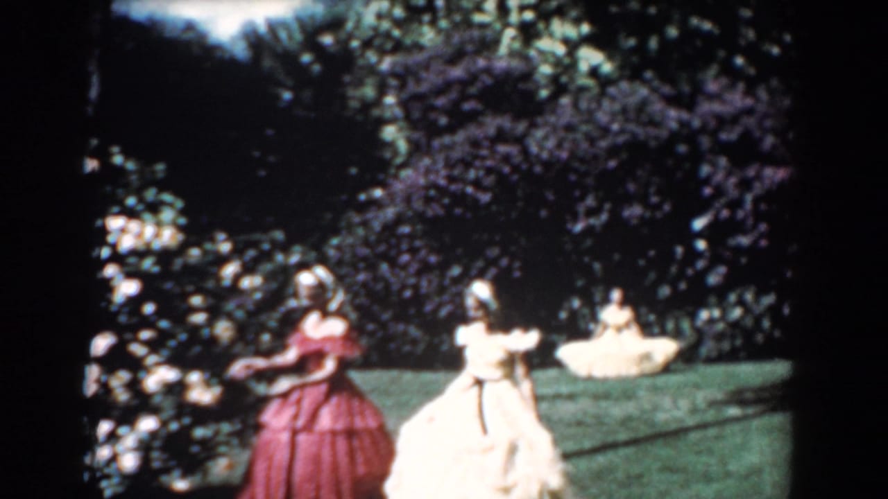 Two women in flowing dresses walk beside rose bushes while another woman sits on the grass behind them.