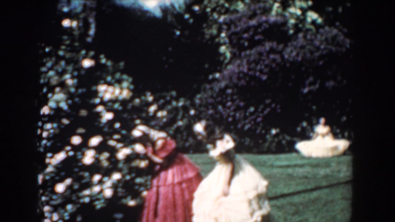 1957 Florida home-movie frame of a woman in red leaning toward roses while another woman in a cream dress watches in the garden.