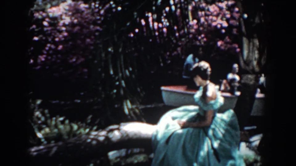 1957 Florida 8mm frame showing a woman in a pale blue gown seated by a shaded riverbank with flowers and a small boat nearby.