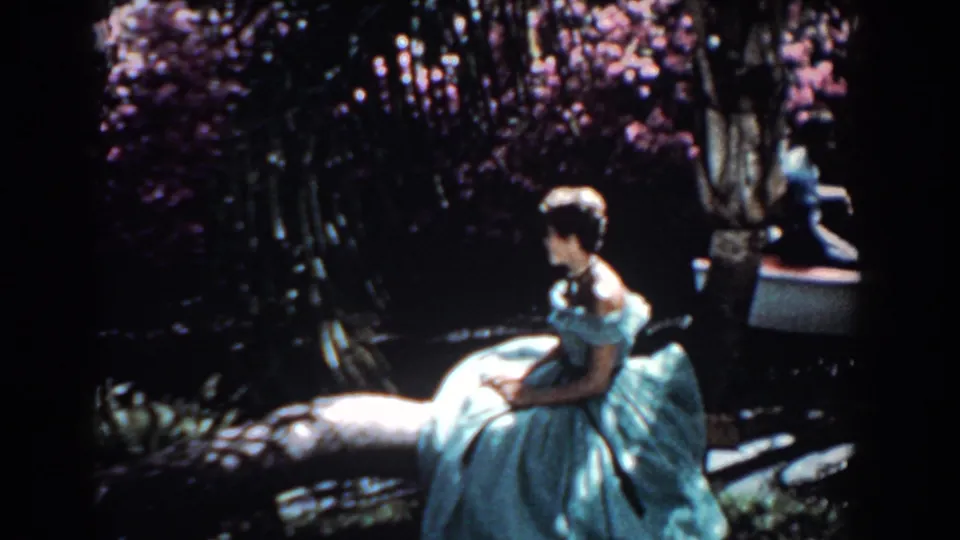 1957 Florida home-movie frame with a woman in a blue gown seated near water, greenery, flowers, and a small boat behind her.