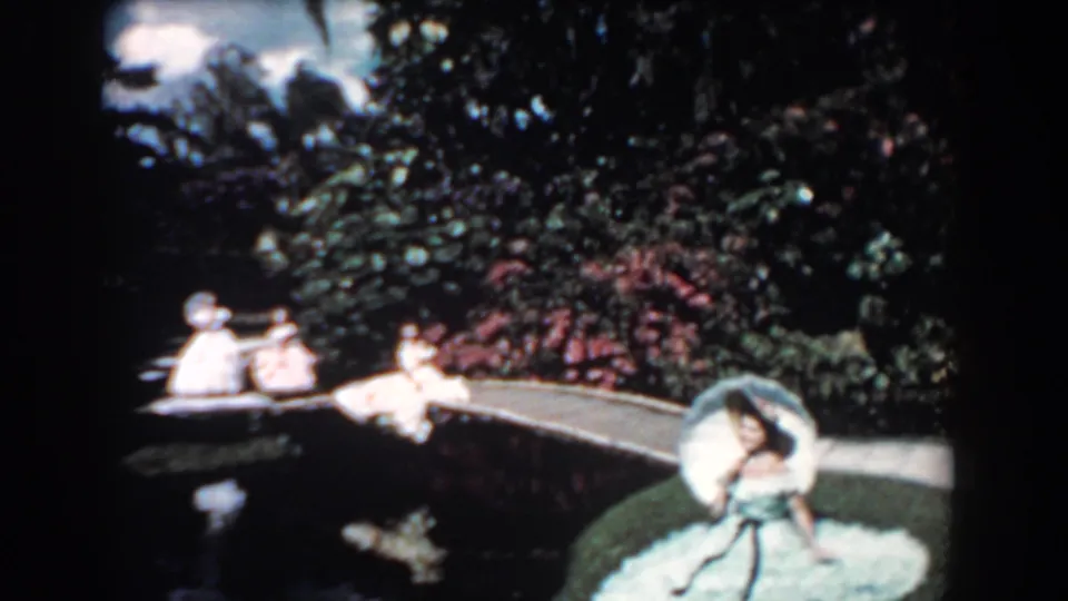 1957 Florida 8mm frame showing a wide riverbank view with people in a small boat, a woman in blue at right, greenery, and a garden path.
