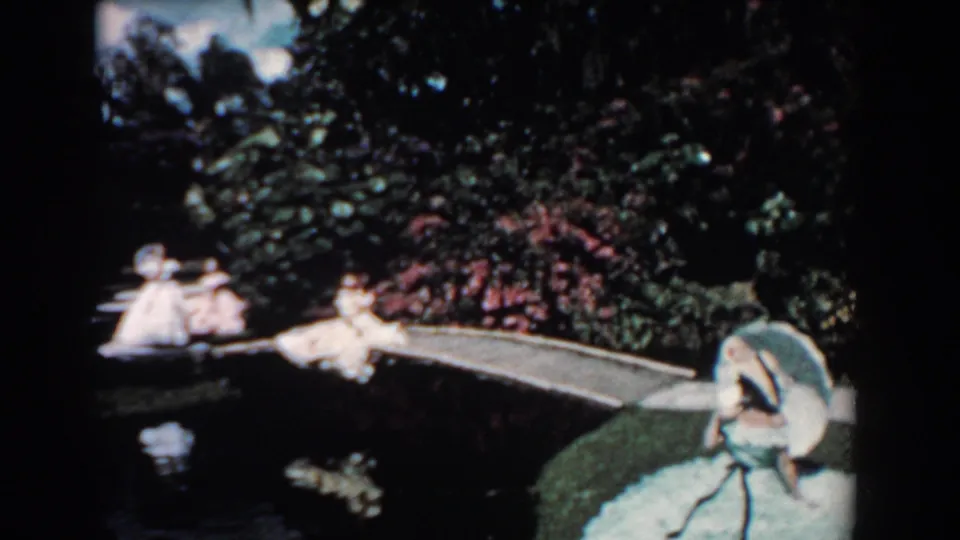 1957 Florida home-movie frame with boat passengers on the left, a woman in a blue gown at right, dense greenery, water, and a path.
