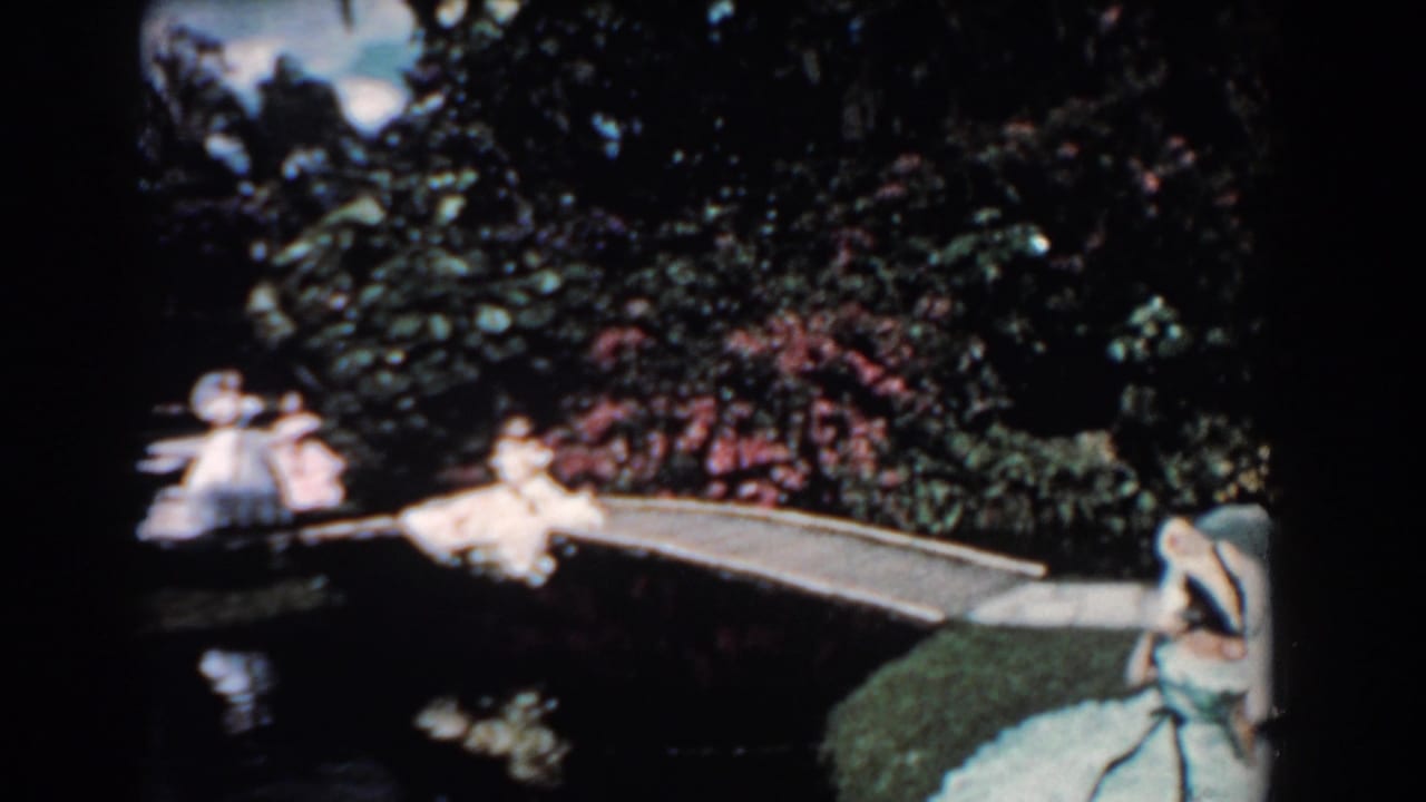 1957 Florida home-movie frame showing women in light dresses and umbrellas around a small garden bridge over a pond.