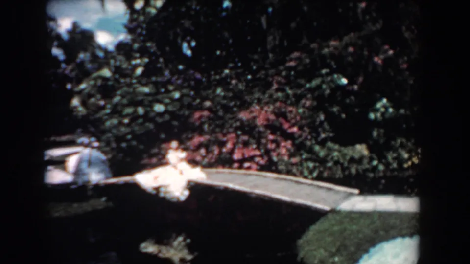 1957 Florida frame with a woman in a light dress on a small bridge, framed by flowering shrubs and pond water.