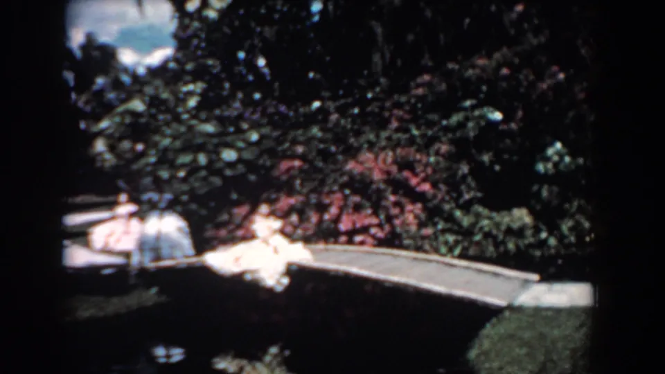 1957 Florida home-movie frame showing women in vintage light dresses with umbrellas near a pond bridge.