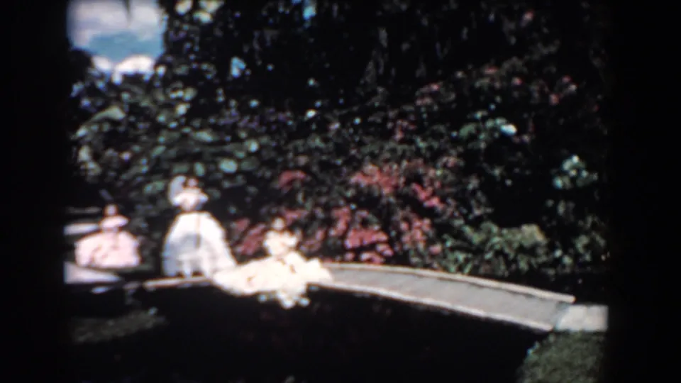 1957 Florida frame with several women in formal light dresses on a small arched bridge amid dense garden foliage.