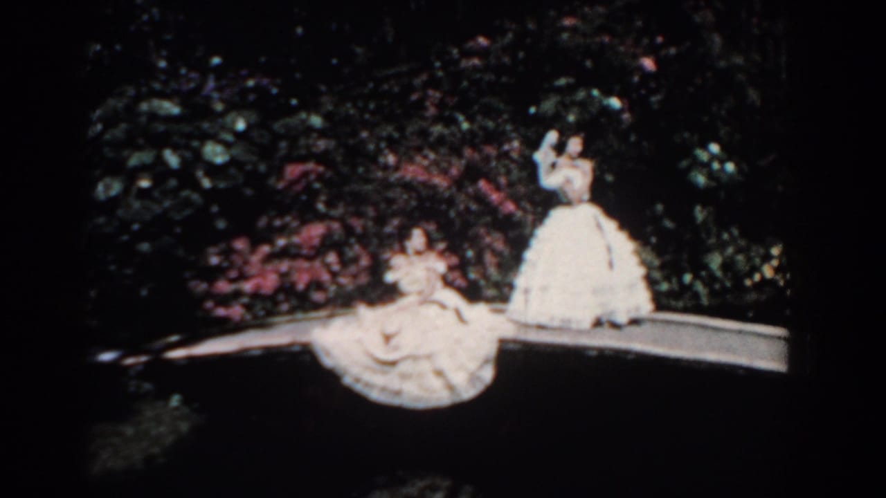 1957 Florida home-movie frame showing two women in full white dresses posed on a garden path amid dense foliage and pink flowering bushes.