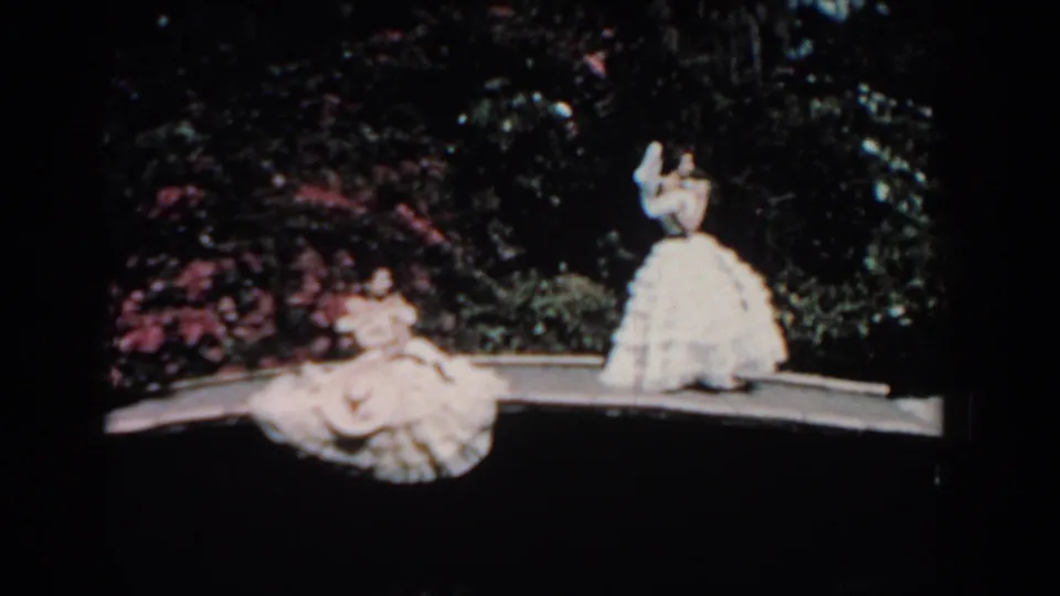 1957 Florida frame with two women in voluminous light dresses on a curved path, backed by dark garden foliage and pink blossoms.