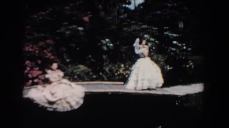 1957 Florida home-movie frame showing two women in white dresses near a garden path edge, with pink flowers and dark foliage in the background.