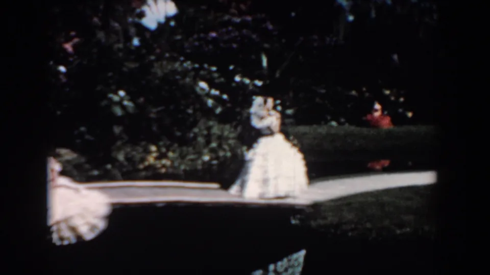 1957 Florida frame with one woman in a full white dress on the curved garden path and a second woman partially visible at the left edge.