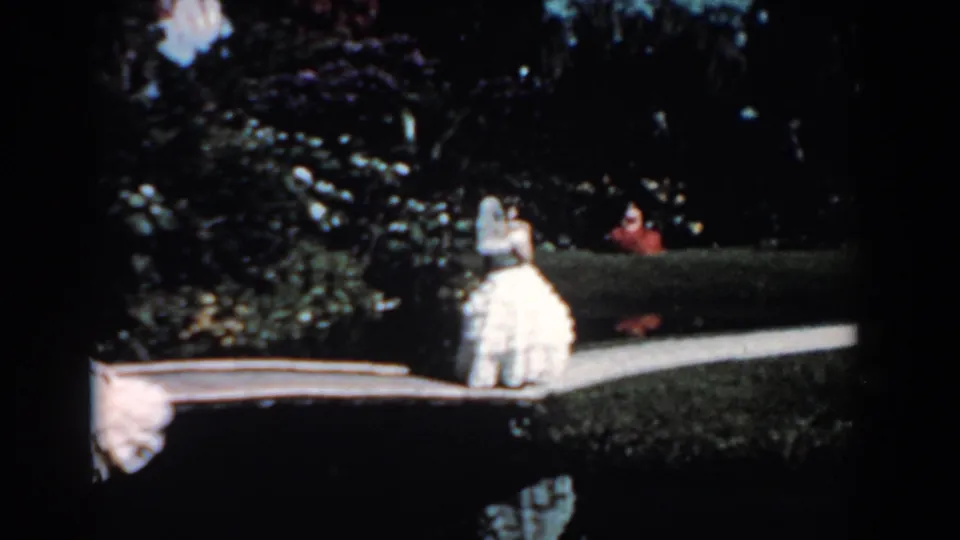 In 1957 Florida home-movie footage, a woman in a white gown appears near a boardwalk bordered by dense greenery.