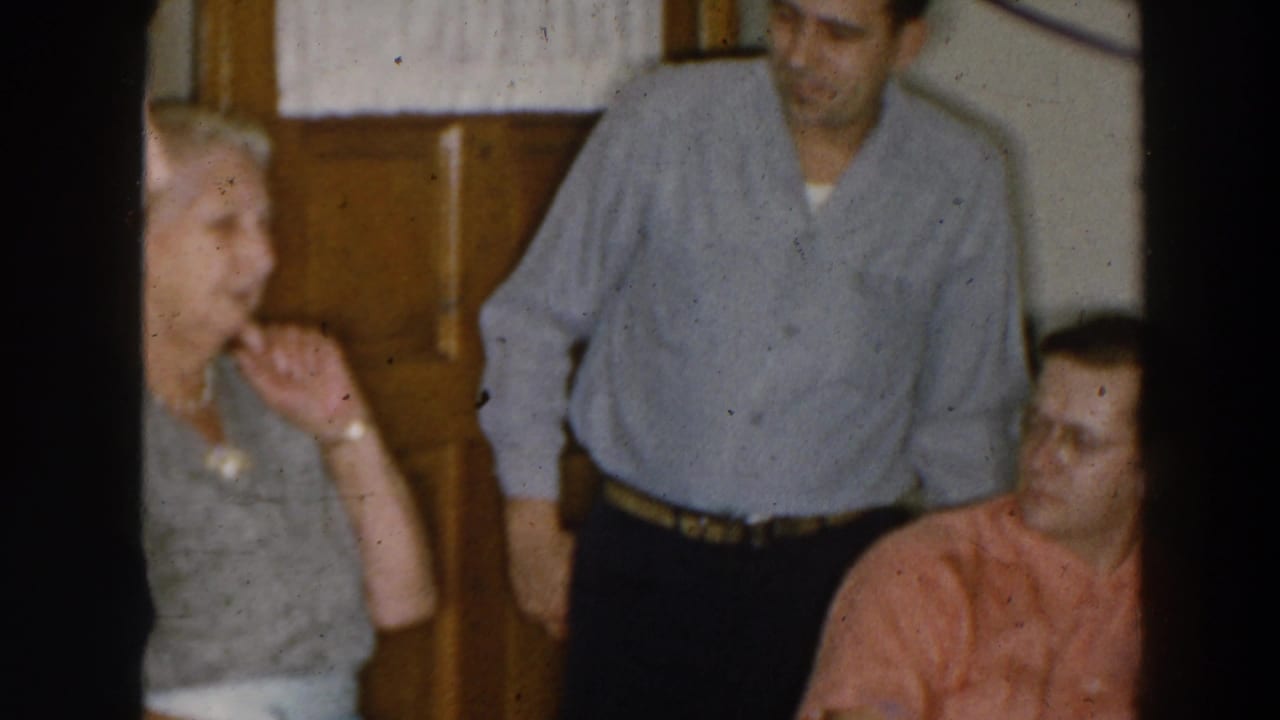Three adults in a home interior, with a woman gesturing beside two men near a wood door.