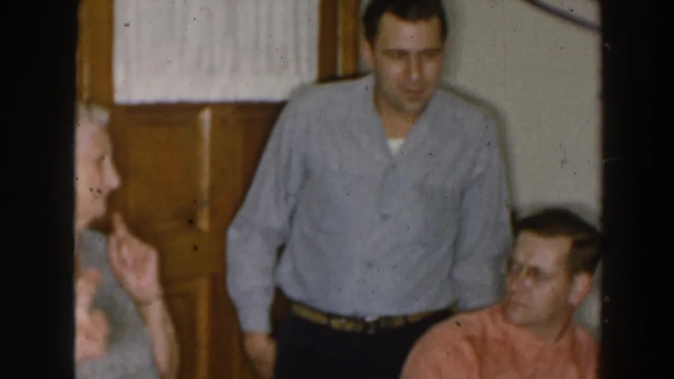 1957 Wheeling Ohio interior frame with a woman at the edge of view speaking to two men in casual shirts.
