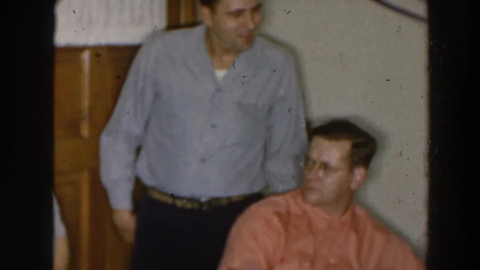 1957 Wheeling Ohio home-movie frame showing a standing man smiling near a seated man in a private room.