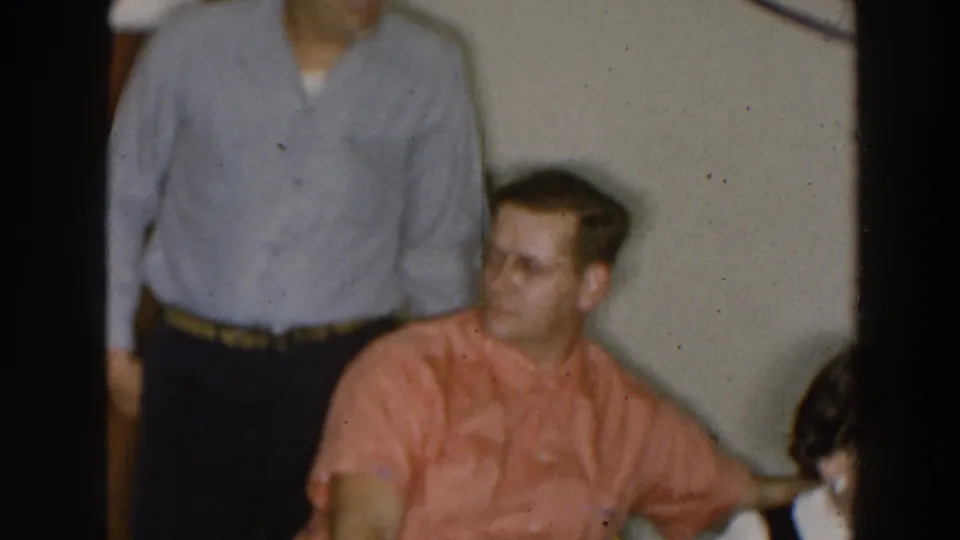 1957 Wheeling Ohio interior frame with a seated man turning during conversation and another man partly visible.